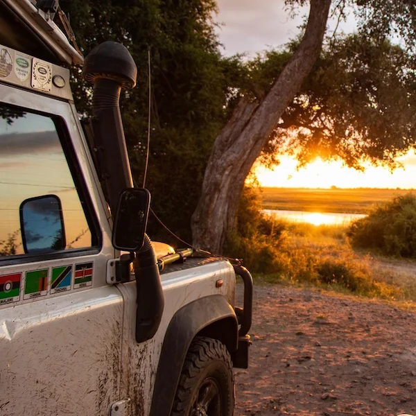 A dusty safari vehicle parked in the African savanna, overlooking a sunset reflecting on a river, with regional flag stickers on its side