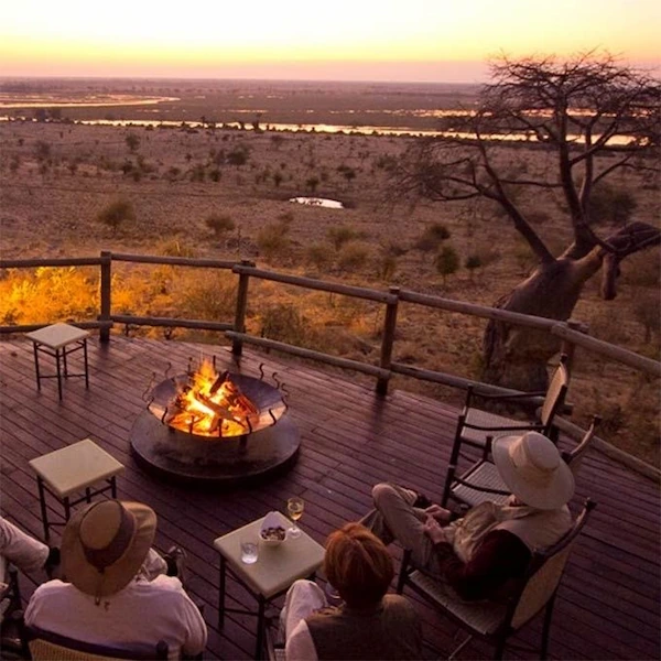 A group of travelers enjoying a campfire on a wooden deck overlooking a vast African savanna at sunset